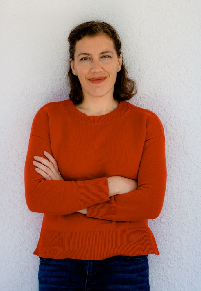 Portrait of Sarah Dzida standing with arms crossed, smiling, wearing an orange sweater.